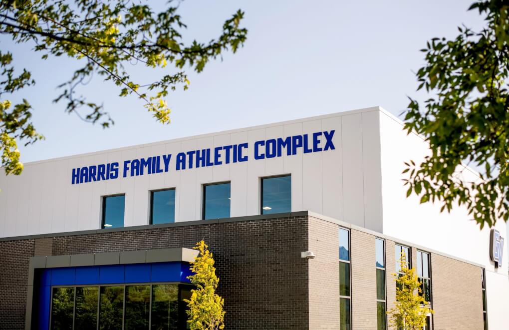 The top part of the white exterior of the Harris Family Athletic Complex is shown. Tree branches frame the image. The words 'Harris Family Athletic Complex' are on the building in GV Blue. 